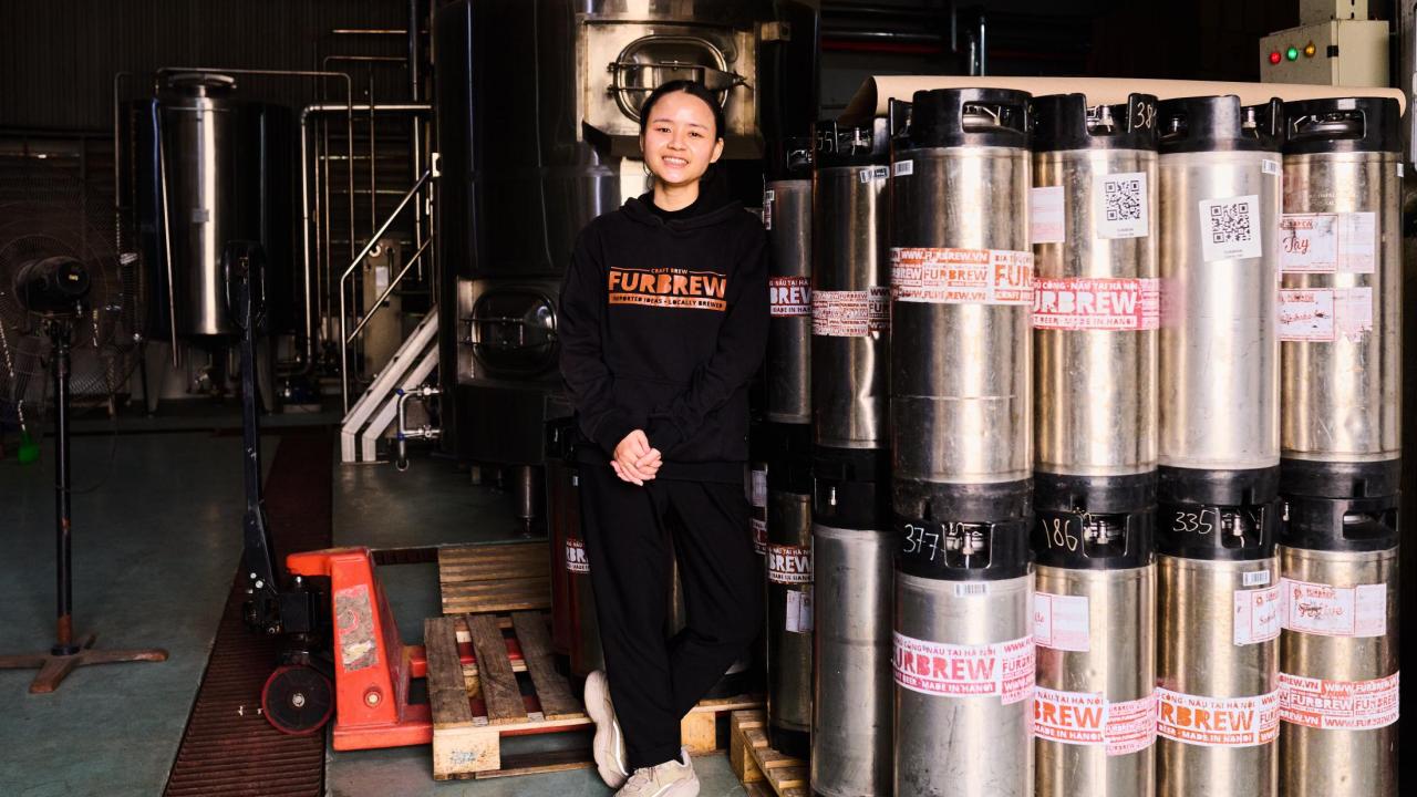 Tapping Potential scholarship winner Dung Ngô stands alongside canisters of beer and other brewing equipment. She wears a black sweatshirt  that reads "Furbrew" in orange lettering.