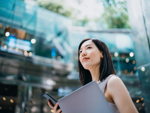 Woman standing in business building atrium. She holds a laptop and looks into the distance.