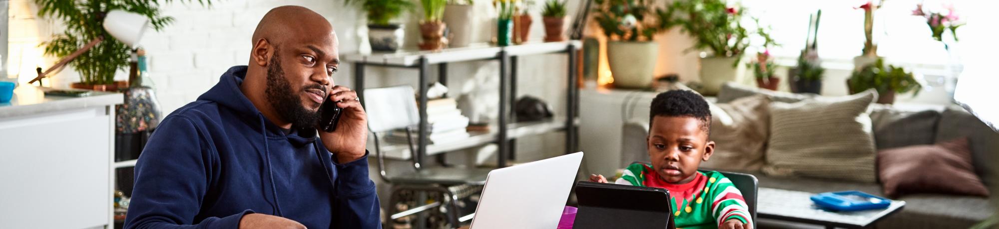 Man working at a laptop at his kitchen table, while his small child plays on a tablet nearby.