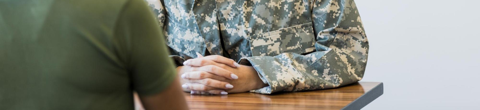 A female military veteran wearing fatigues sits across a table from a woman in civilian clothes